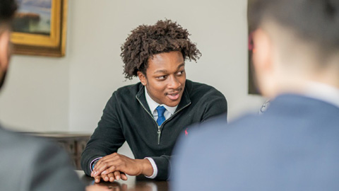 A person wearing a dark sweater and blue tie, sits at a table.
